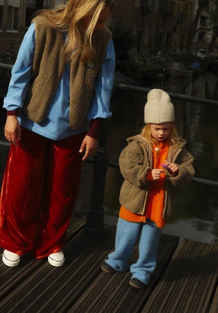 Woman and child in knit hats and warm clothes standing on a wooden bridge over a canal in an urban area on a sunny day.