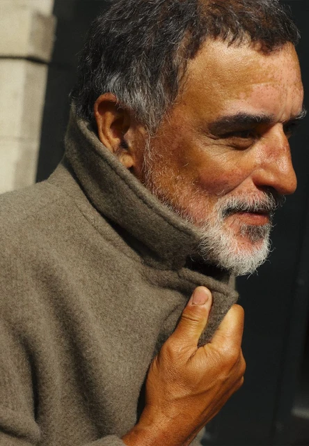 Close-up portrait of a middle-aged man with a grey beard wearing a tan wool coat, looking off to the side with closed eyes.
