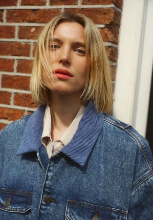 Portrait of a blonde woman wearing a denim jacket with blue corduroy collar against a brick wall