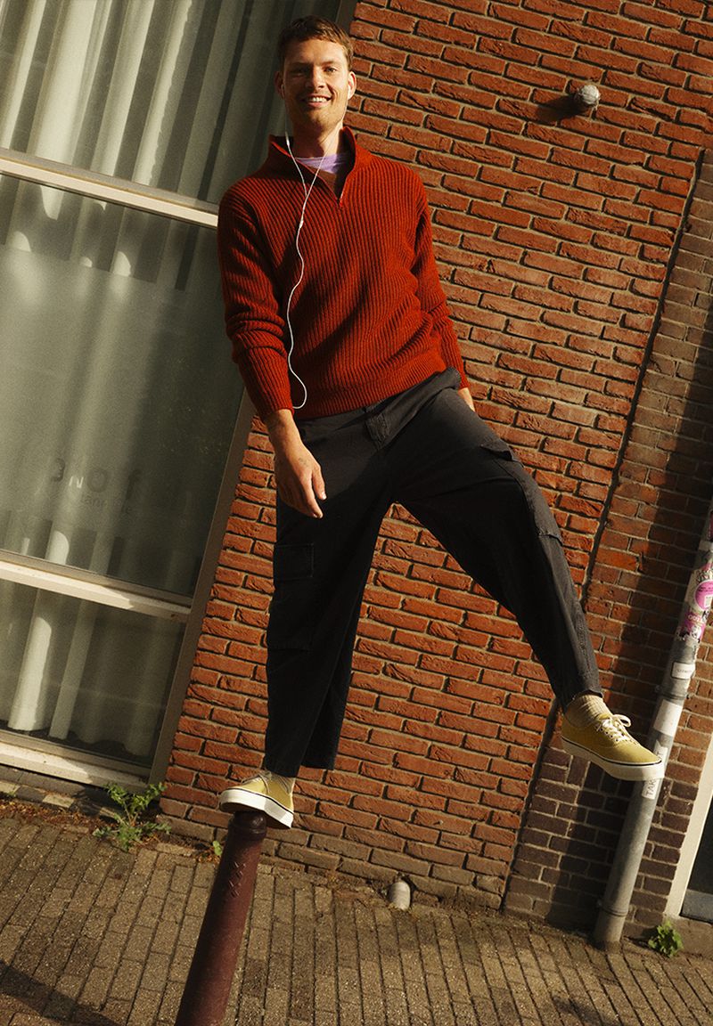 Young adult male standing on a metal pole in an outdoor urban environment with a brick wall and window in the background.