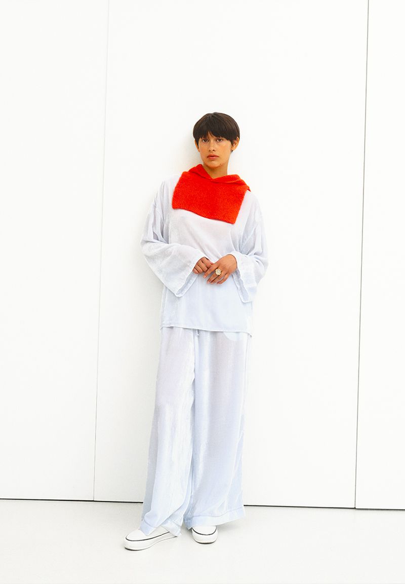 A woman stands in a photo studio wearing an oversized white outfit and sneakers, with a bright red scarf. The background is plain white.