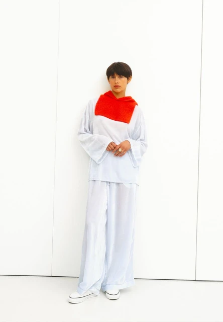 A woman stands in a photo studio wearing an oversized white outfit and sneakers, with a bright red scarf. The background is plain white.