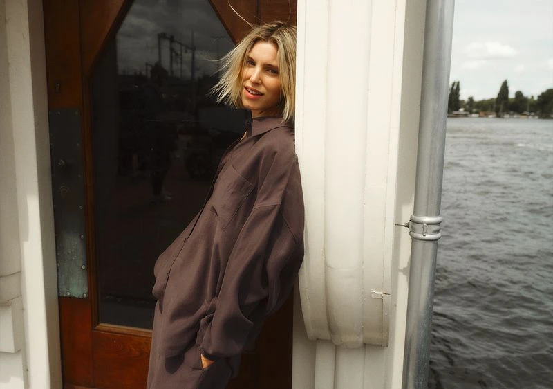Blonde woman in a brown shirt dress leans against a white column on a boat, with a river and trees visible in the background.