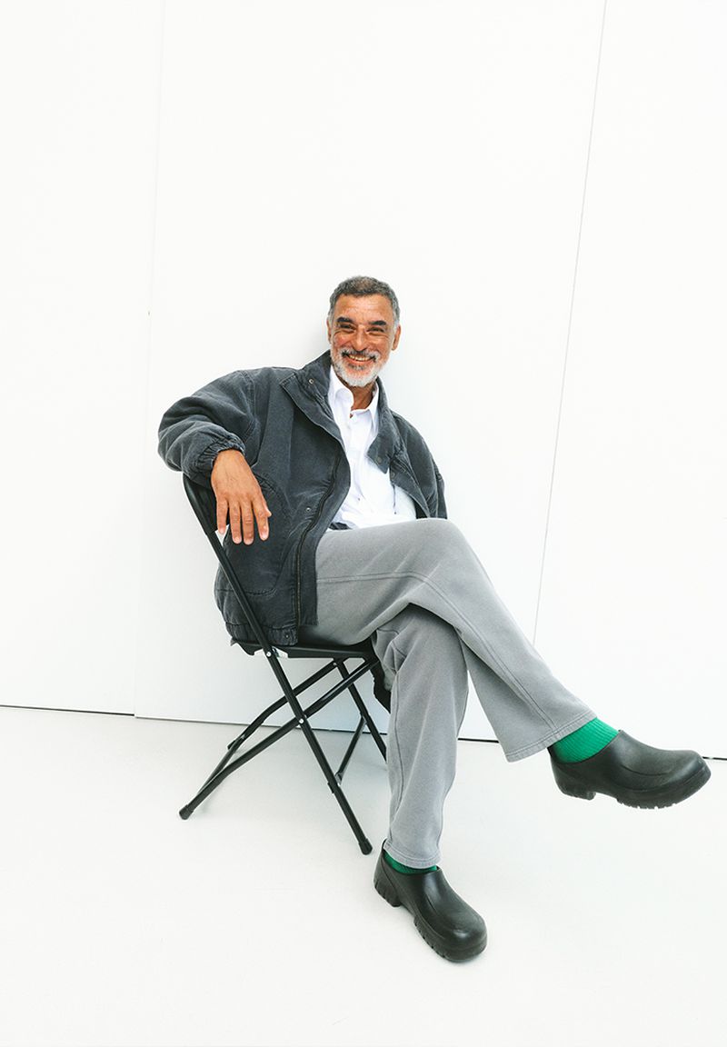 Man in a jacket, seated, smiling against a white wall. He wears grey pants and black shoes with green socks, creating a casual, inviting scene.