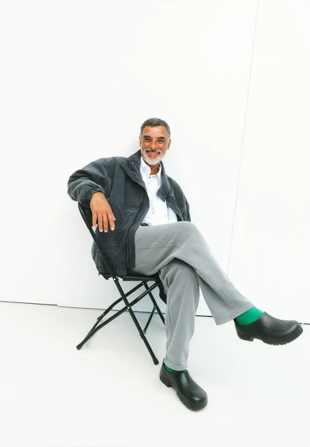 Man in a jacket, seated, smiling against a white wall. He wears grey pants and black shoes with green socks, creating a casual, inviting scene.