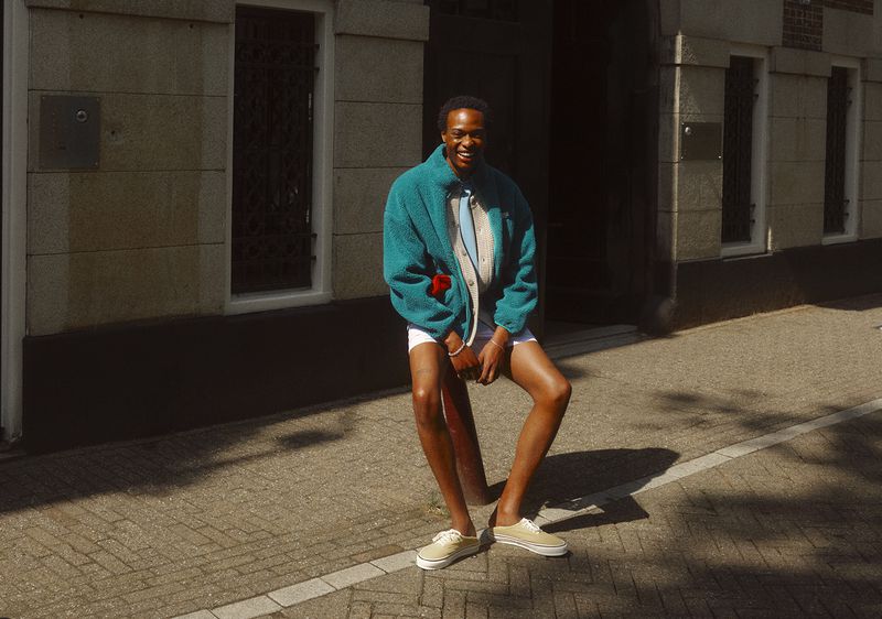 A person in a turquoise fleece jacket and tan sneakers poses outside a stone building on a sunny day, casting shadows on the sidewalk.
