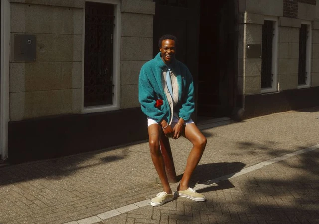 A person in a turquoise fleece jacket and tan sneakers poses outside a stone building on a sunny day, casting shadows on the sidewalk.