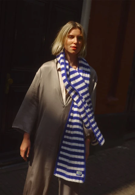 Fashion shot of a woman wearing a taupe dress and a blue and white striped scarf, standing outdoors on a sunny day.