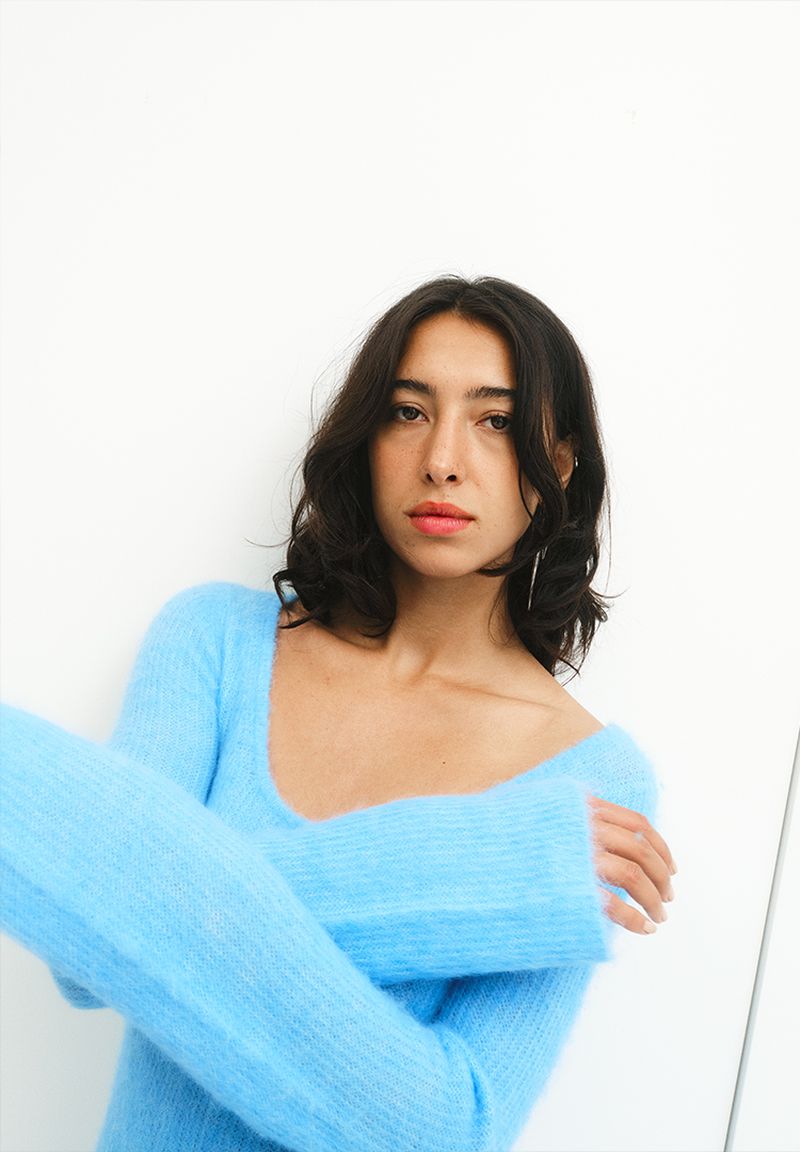 Portrait of a young woman with dark hair wearing a fluffy, light blue sweater against a plain white background, looking at the camera.