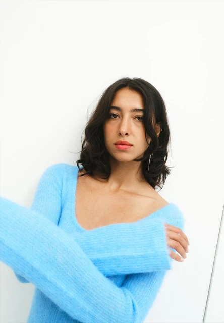 Portrait of a young woman with dark hair wearing a fluffy, light blue sweater against a plain white background, looking at the camera.