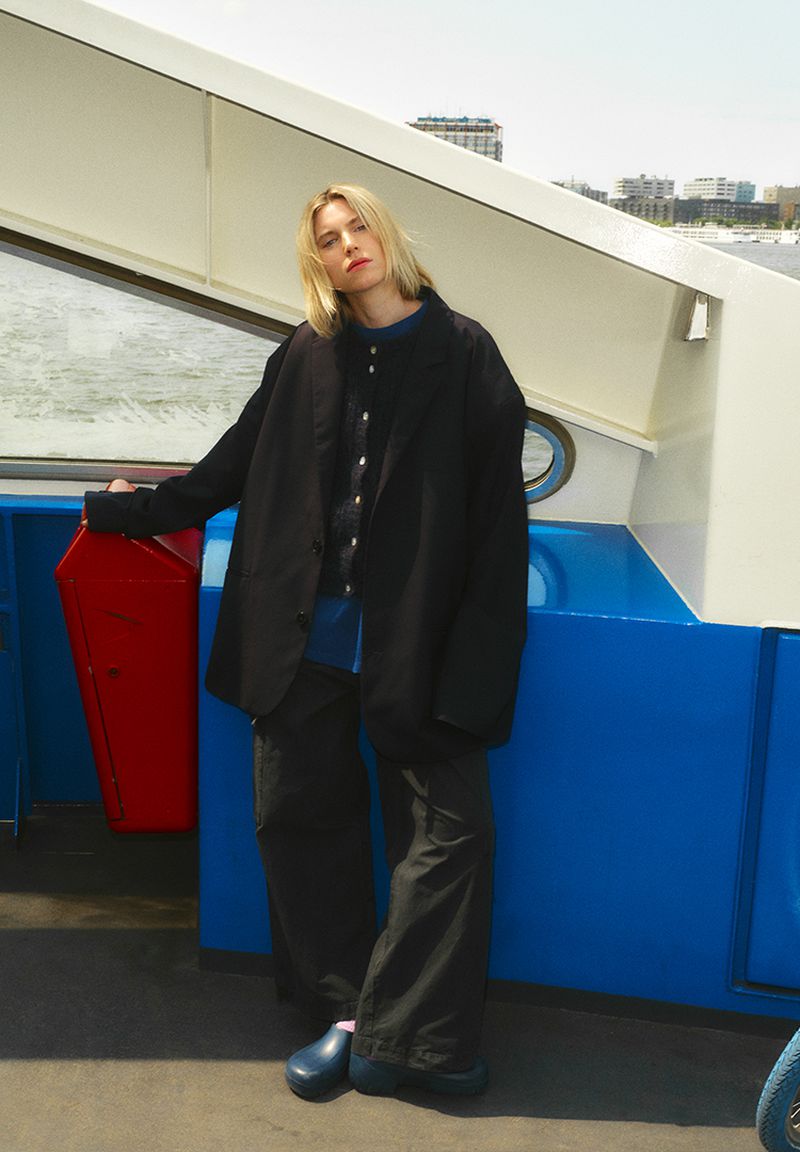 A young adult woman stands on a boat wearing a black jacket, wide leg pants, and clogs on a sunny day with city skyline background.