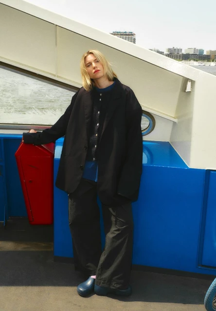 A young adult woman stands on a boat wearing a black jacket, wide leg pants, and clogs on a sunny day with city skyline background.