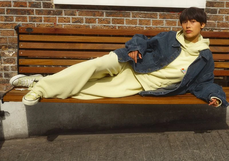 Woman in light yellow sweatsuit and denim jacket lounges on wooden bench with a brick wall background in an outdoor urban setting.