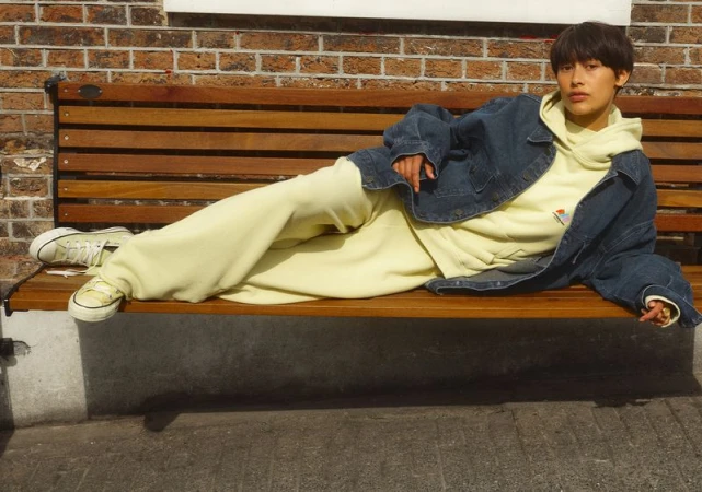 Woman in light yellow sweatsuit and denim jacket lounges on wooden bench with a brick wall background in an outdoor urban setting.