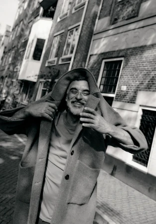 Smiling older man in Amsterdam wearing a hooded coat, photographed by Rasmus Weng Karlsen in black and white.