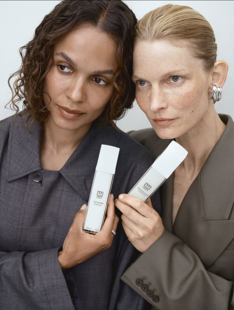 Two women pose with a white skincare product. One woman with tan skin and curly hair looks left, and the other with light skin looks forward.