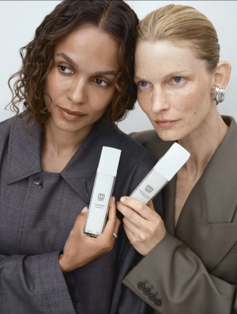 Two women pose with a white skincare product. One woman with tan skin and curly hair looks left, and the other with light skin looks forward.
