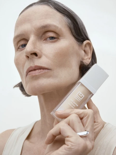 Elegant older adult woman poses in a close up shot, holding a skincare bottle in a beauty product shot, set against a white backdrop.