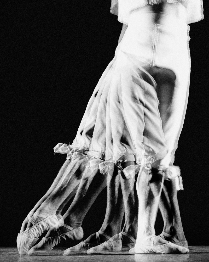 Multiple exposures of a ballet dancer wearing white and ballet slippers performing a series of leg movements against a dark backdrop.