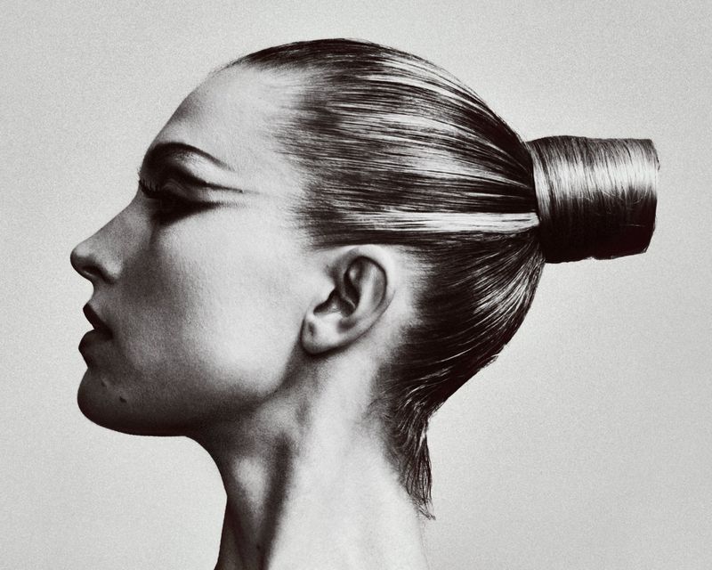 Monochrome portrait of an adult woman in profile with slicked-back hair styled in a high bun and dramatic winged eyeliner.