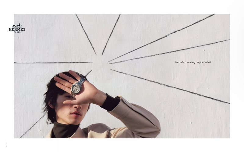 Person with a luxury watch holding hand to the face standing against an off-white wall with hand-drawn sunburst design, outdoors.
