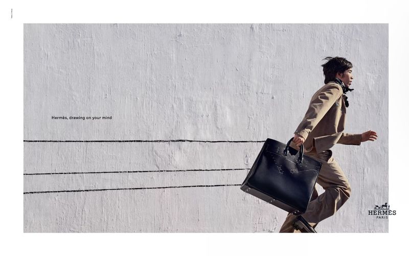 A man in a tan suit runs with a black bag against a white wall in a fashion advertisement with graphic black lines and text.