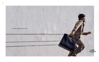 Model running along a white textured wall carrying a large black leather bag for Hermes