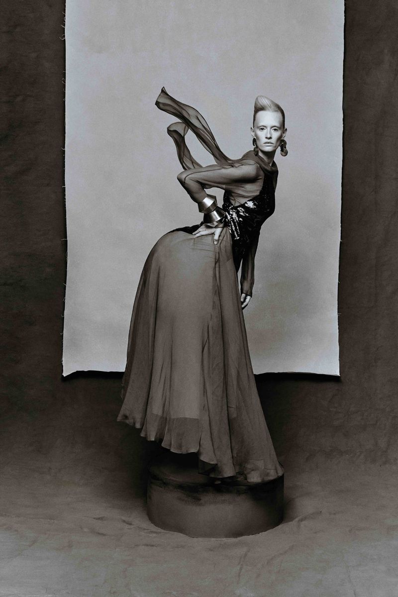 Monochrome portrait of a female model wearing a long sheer gown, statement earrings, and a metal cuff bracelet in a studio setting.
