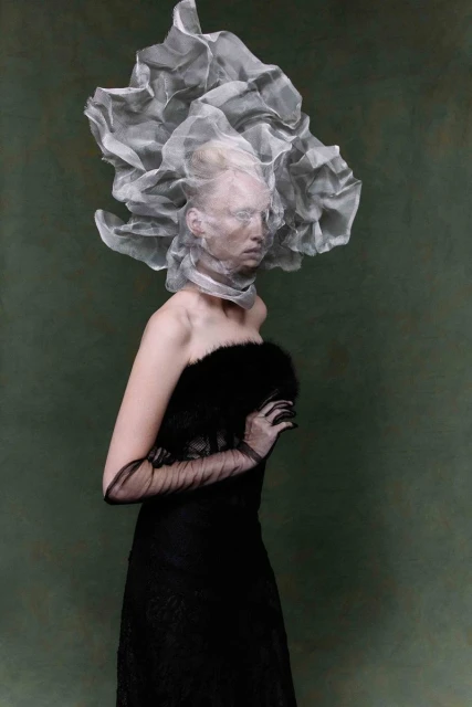 Elegant woman in a black dress, sheer gloves, and a ruffled white headpiece against an olive green background in a fashion style photograph.