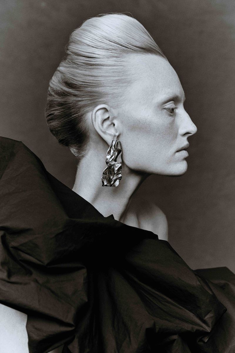 Elegant woman with platinum blonde updo and large silver earrings. She is wearing a dark flowing garment against a grey background.