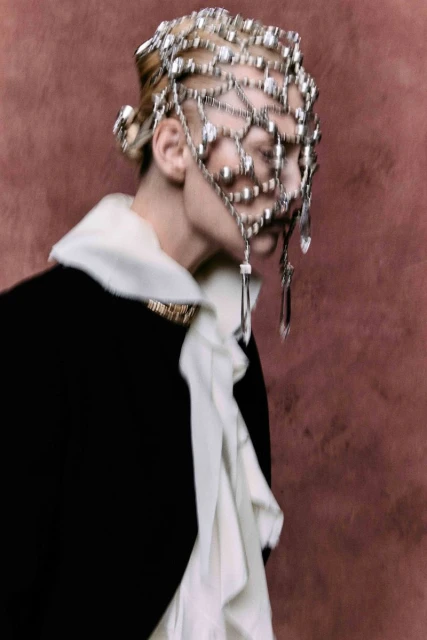 Fashion portrait of a person wearing a jeweled head covering, black jacket with a white ruffled collar, and gold brooch against a mauve background.