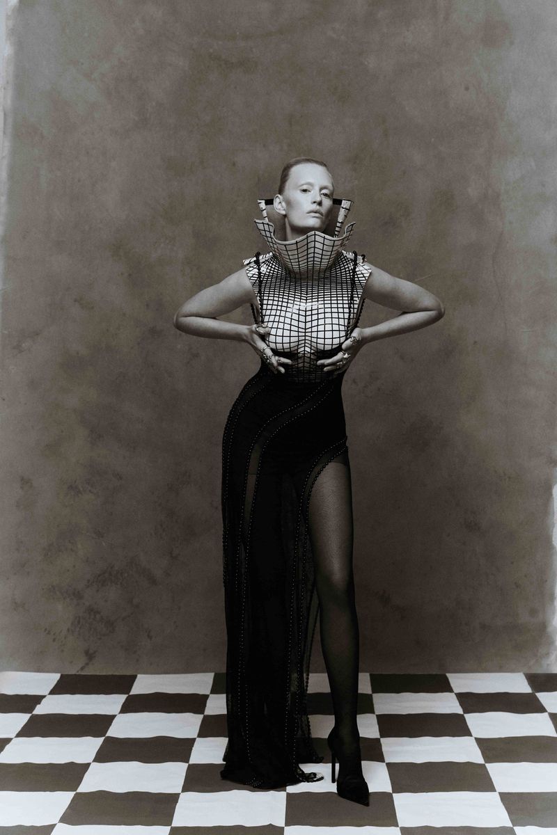 Elegant woman in a structural grid-patterned top and a long black dress with a thigh-high slit poses on a black-and-white checkered floor.