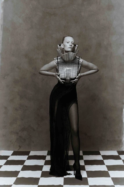 Elegant woman in a structural grid-patterned top and a long black dress with a thigh-high slit poses on a black-and-white checkered floor.