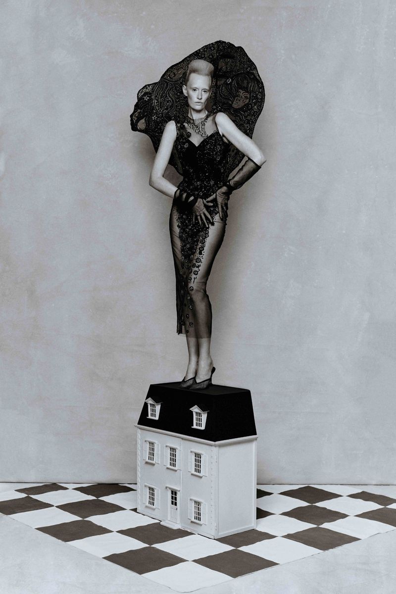 Elegant woman in lace gown stands on a dollhouse, set on a checkered floor against a muted gray wall in a creative fashion photograph.