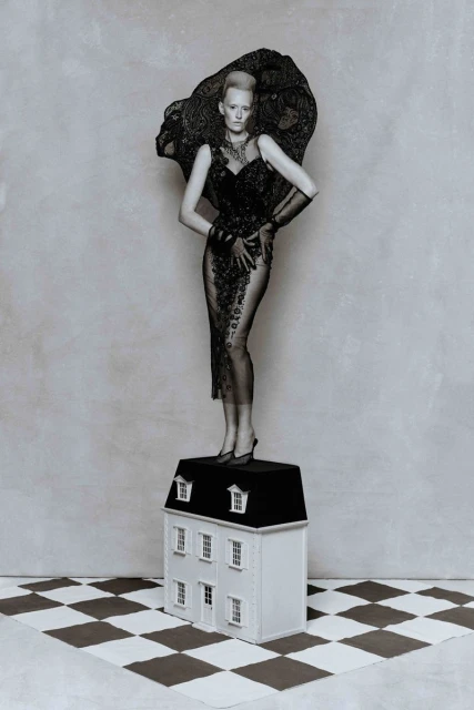 Elegant woman in lace gown stands on a dollhouse, set on a checkered floor against a muted gray wall in a creative fashion photograph.