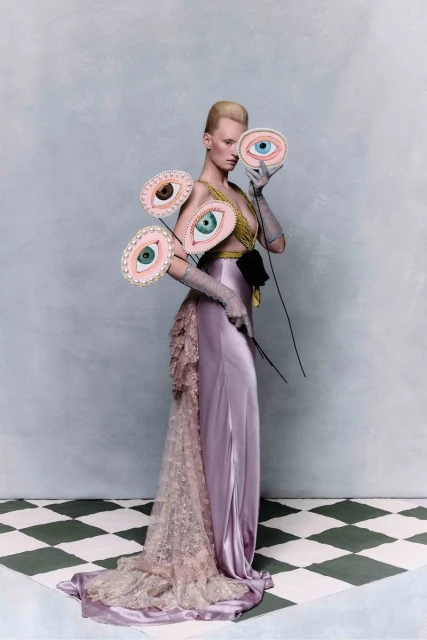 Woman poses in an elaborate lavender gown, holding a decorative eye mask, with more eye masks pinned asymmetrically to her dress.