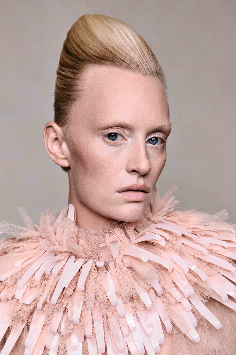 Portrait of a fair-skinned woman with platinum blonde hair in an updo, wearing a feather-like collar, against a muted grey background.