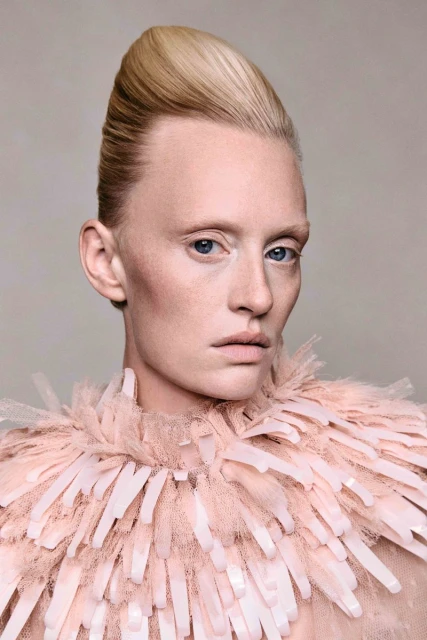 Portrait of a fair-skinned woman with platinum blonde hair in an updo, wearing a feather-like collar, against a muted grey background.