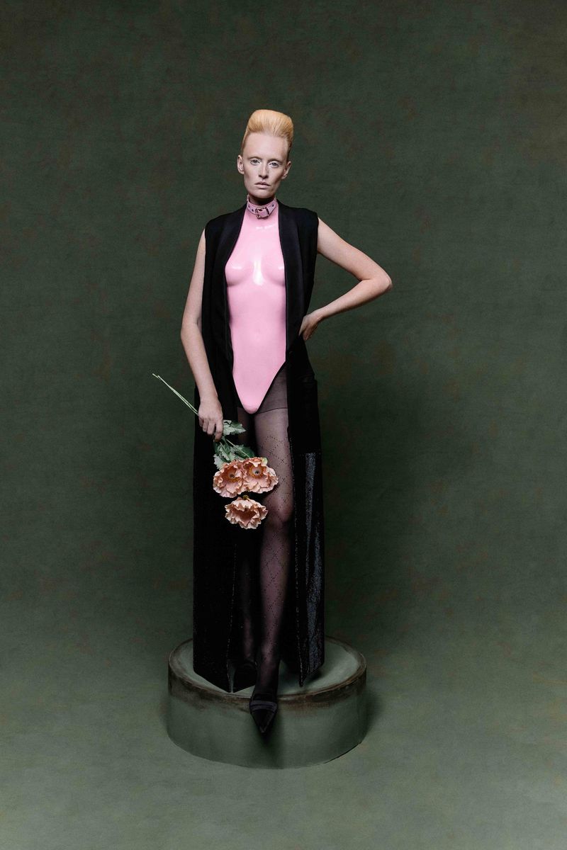 Woman with pale skin and blonde hair stands on a plinth in a pink leotard, black vest, and holds flowers against a green background.