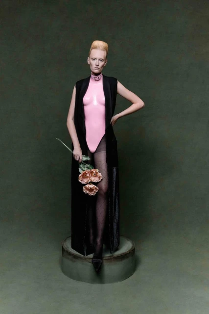 Woman with pale skin and blonde hair stands on a plinth in a pink leotard, black vest, and holds flowers against a green background.