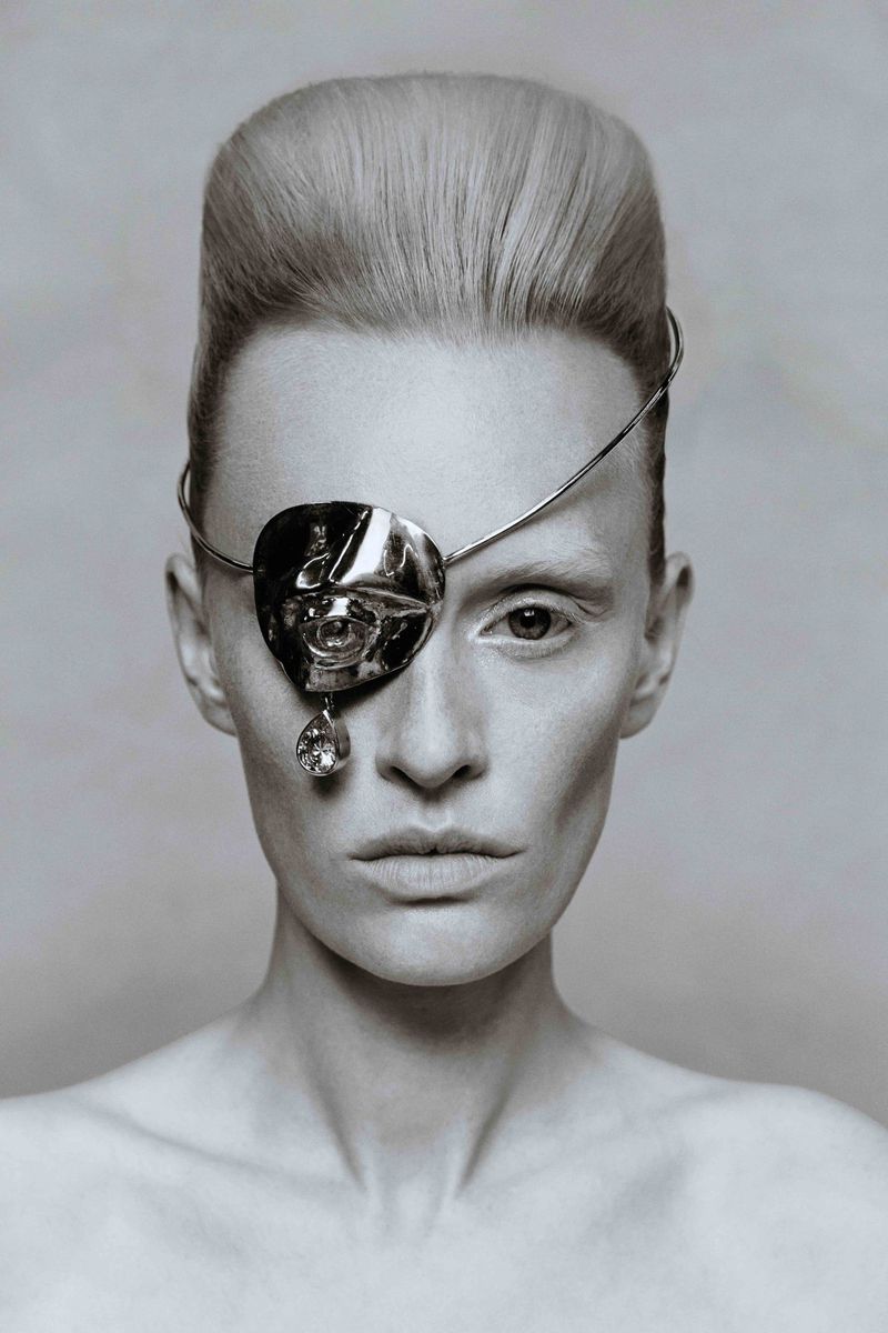 Black-and-white portrait of a fair-skinned adult woman with platinum blonde hair, wearing an ornate silver metal eyepatch and blank expression.