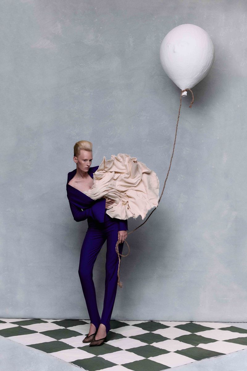 Fashion shot of an adult female in a dark purple suit, holding a large, light beige, ruffled fabric, and a white balloon on a string.
