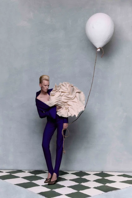 Fashion shot of an adult female in a dark purple suit, holding a large, light beige, ruffled fabric, and a white balloon on a string.