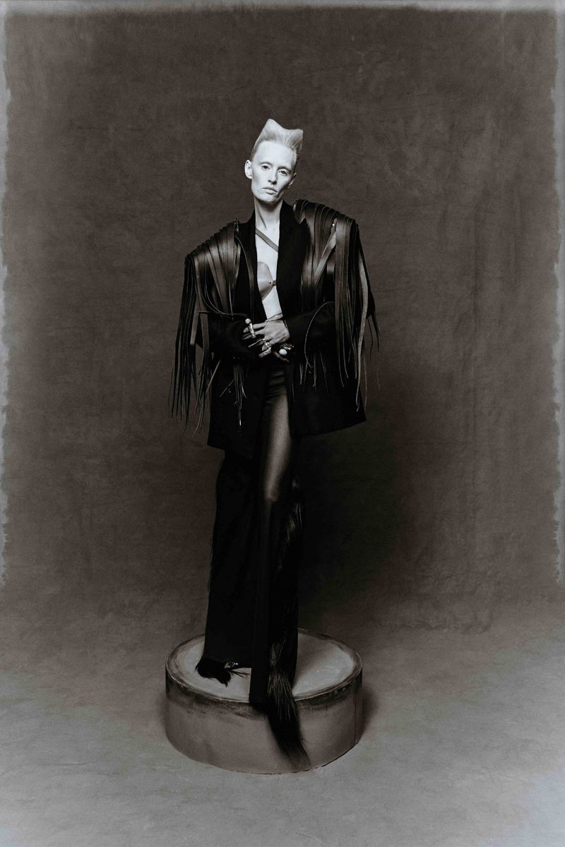 Monochrome portrait of a slender adult female with a short, styled platinum hairdo wearing a black fringed jacket and standing on a cylinder.