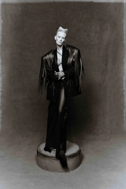 Monochrome portrait of a slender adult female with a short, styled platinum hairdo wearing a black fringed jacket and standing on a cylinder.