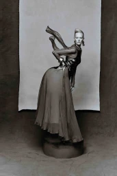 Model Luca Aimee poses on a low pedestal in a flowing sheer dress with an airborne scarf against a studio backdrop.