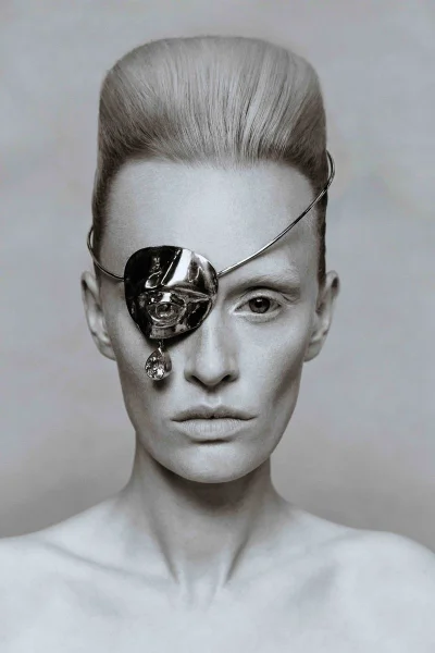 Close-up portrait of a model wearing a sculptural metallic eye patch with a dangling gem and structured beehive hairstyle.