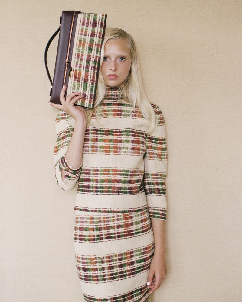 Woman posing indoors with a brown leather and plaid handbag. She is wearing a plaid dress with shades of orange and green.
