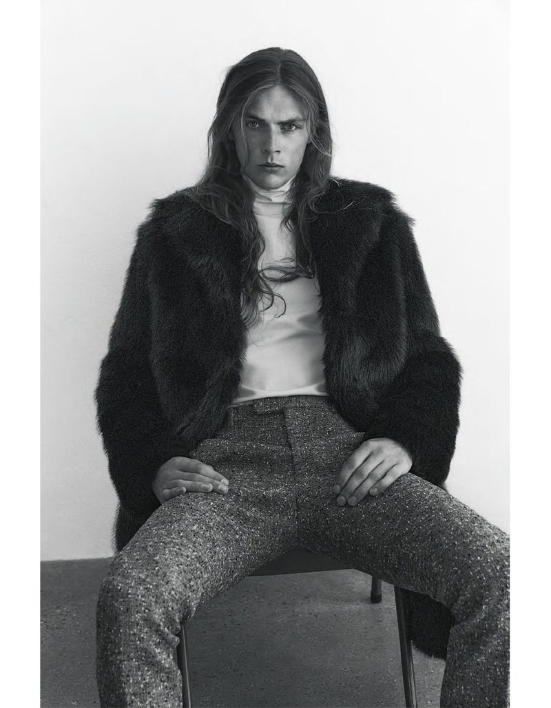 Monochrome portrait of a long-haired male wearing a fur coat and trousers while sitting on a chair in a photo studio.