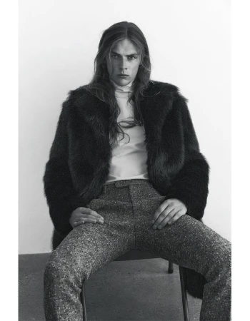 Monochrome portrait of a long-haired male wearing a fur coat and trousers while sitting on a chair in a photo studio.